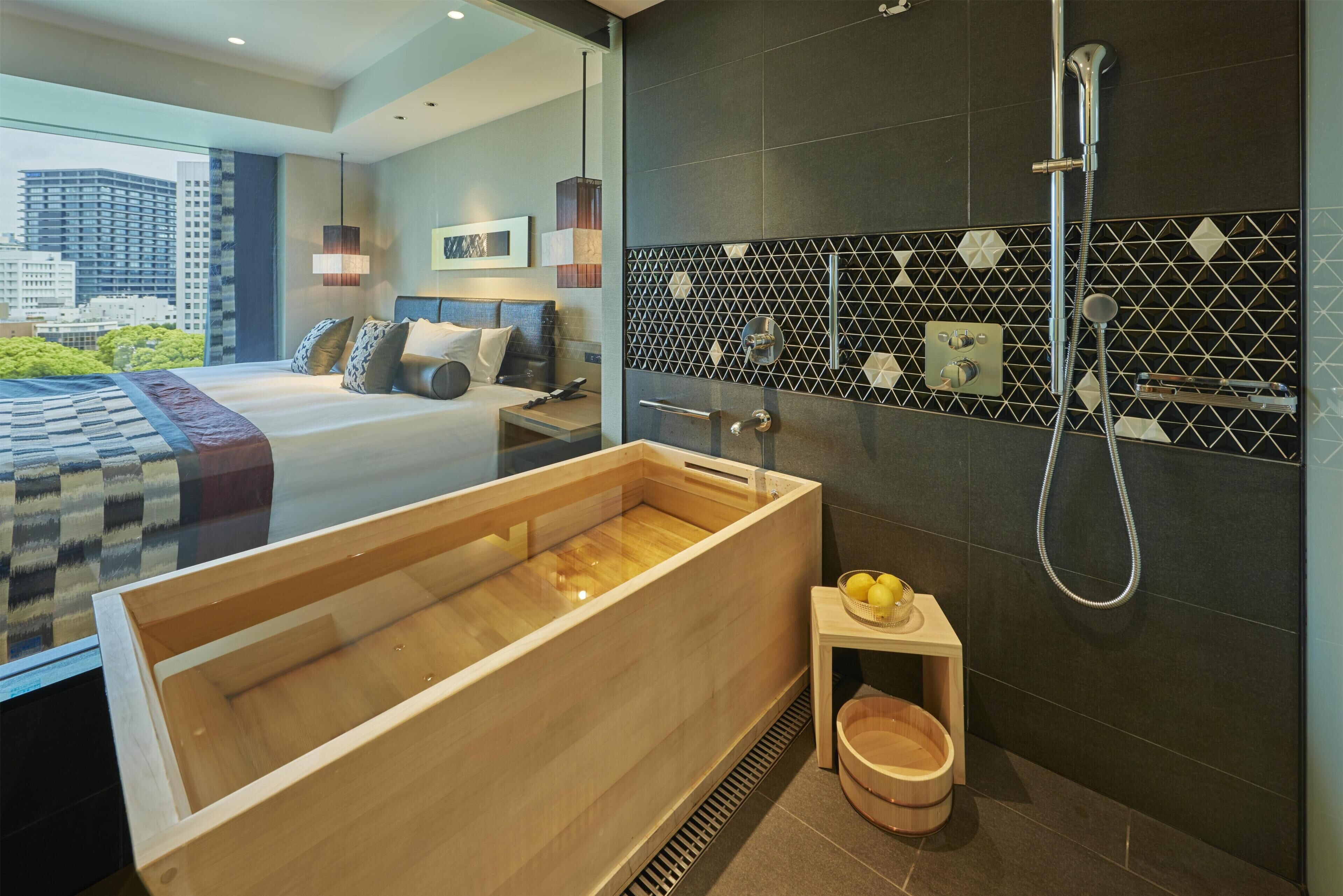 A modern hotel room with a large wooden bathtub, a shower, a small table with a basket, and a bed with a city view.