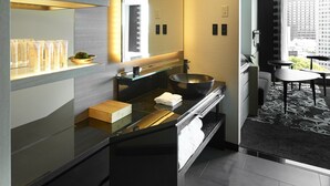 A modern bathroom with a large mirror, a black countertop, and a bowl sink. There is a shelf with toiletries and a small plant. The floor is tiled, and there is a window with a view of buildings.