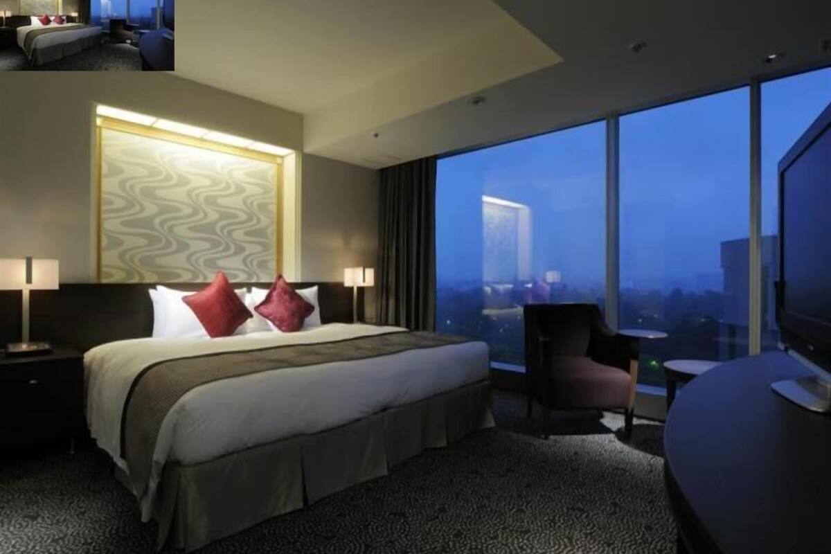 A hotel room with a large bed, two bedside tables, a chair, a TV, and a large window with a city view.