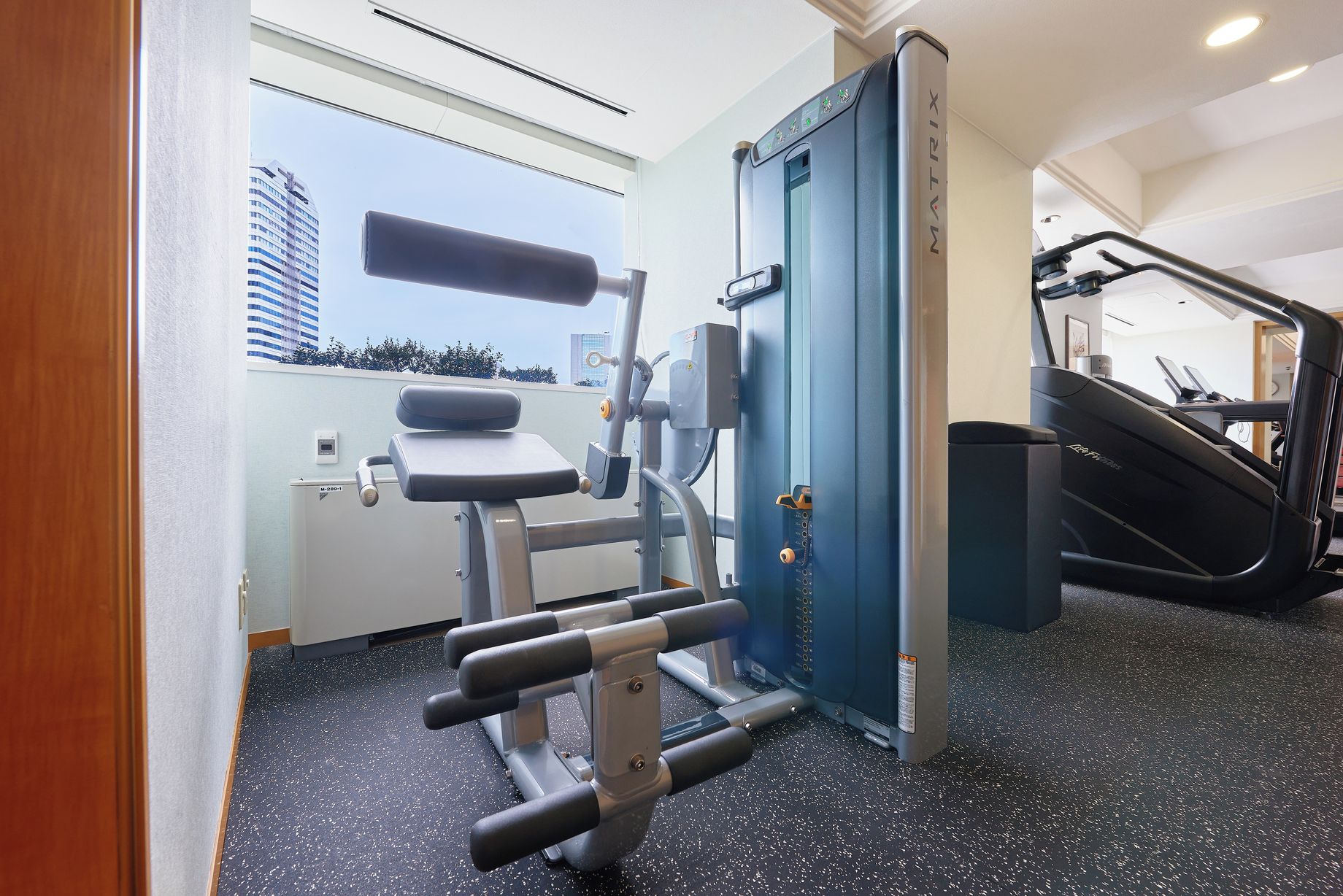 Une salle de sport équipée de matériel d’exercice, d’un tapis de course et offrant une vue sur un bâtiment à l’extérieur.