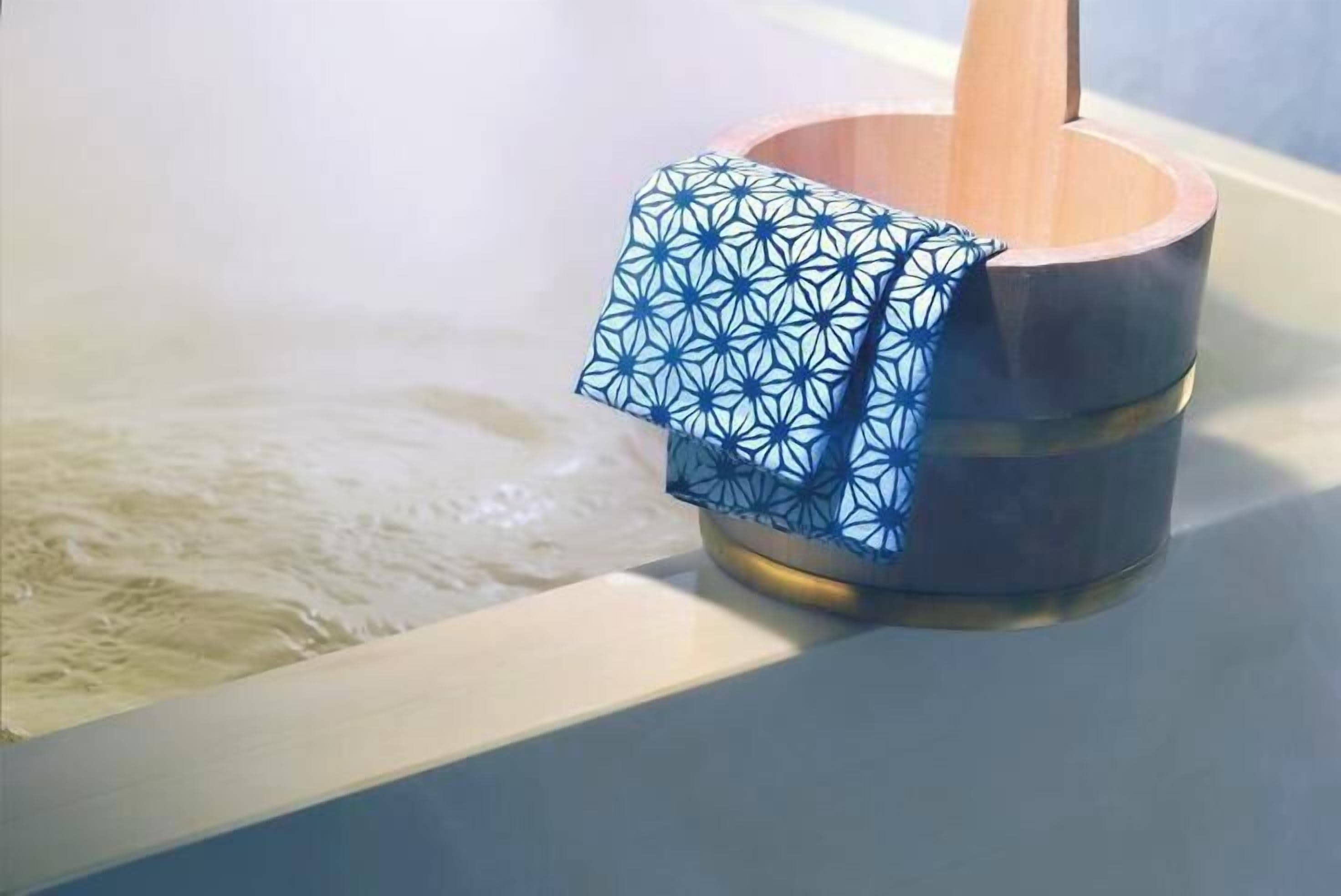 Un balde de madera con un paño azul estampado en su interior, sobre una superficie blanca, junto a una bañera con agua.