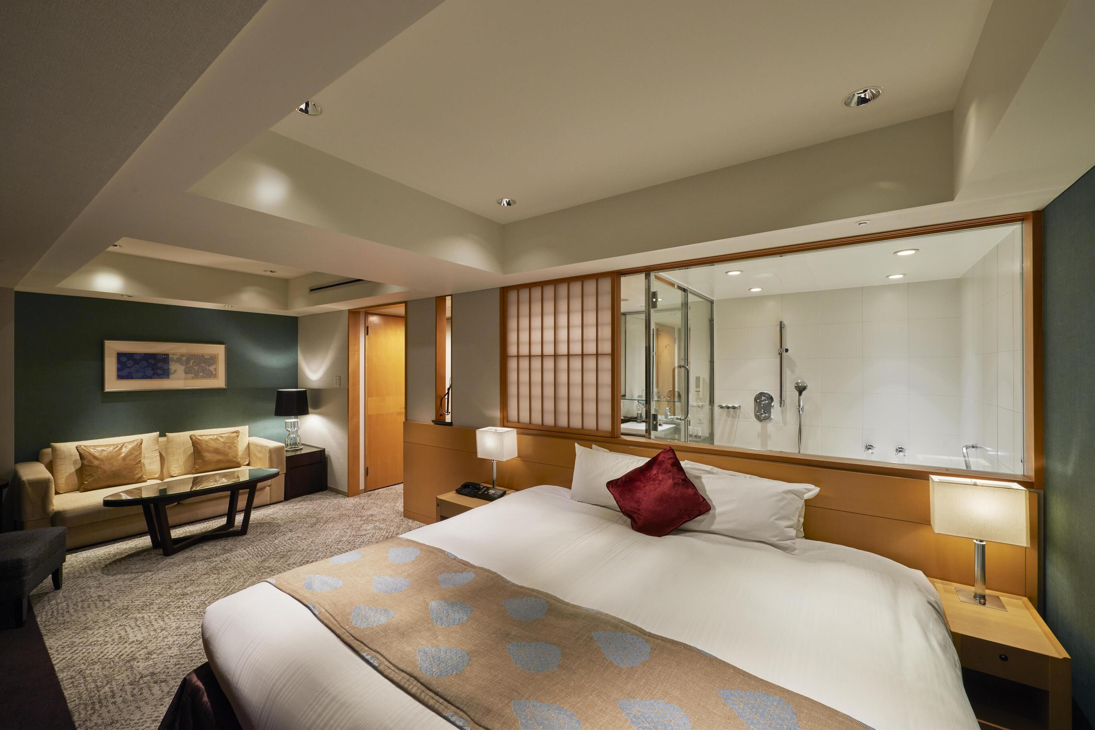 A hotel room with a large bed, a sofa, a desk, and a bathroom with a glass shower.