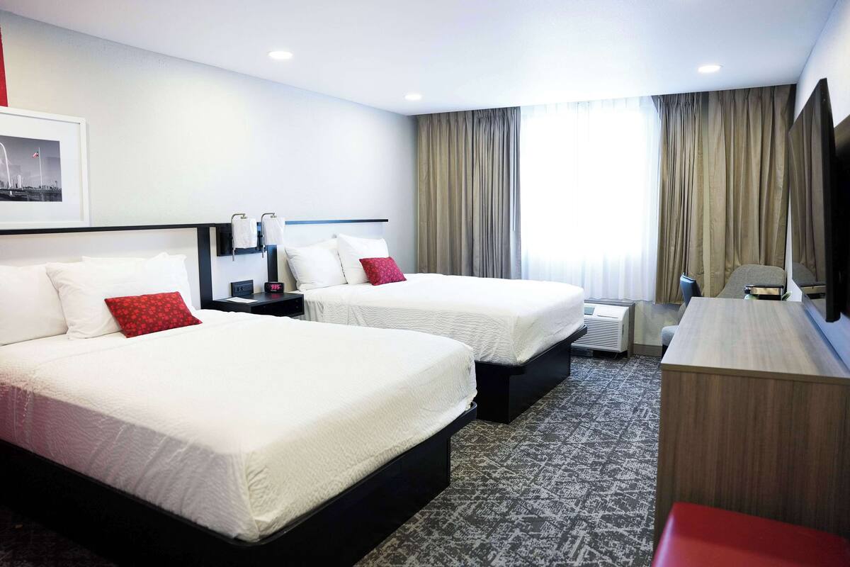 A hotel room with two beds, a desk, a TV, and a large window with curtains.