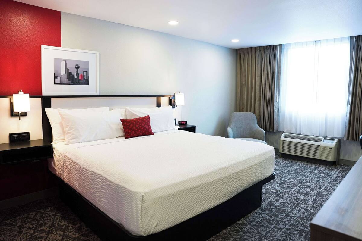 A hotel room with a large bed, a cityscape picture on the wall, a chair, and an air conditioning unit.