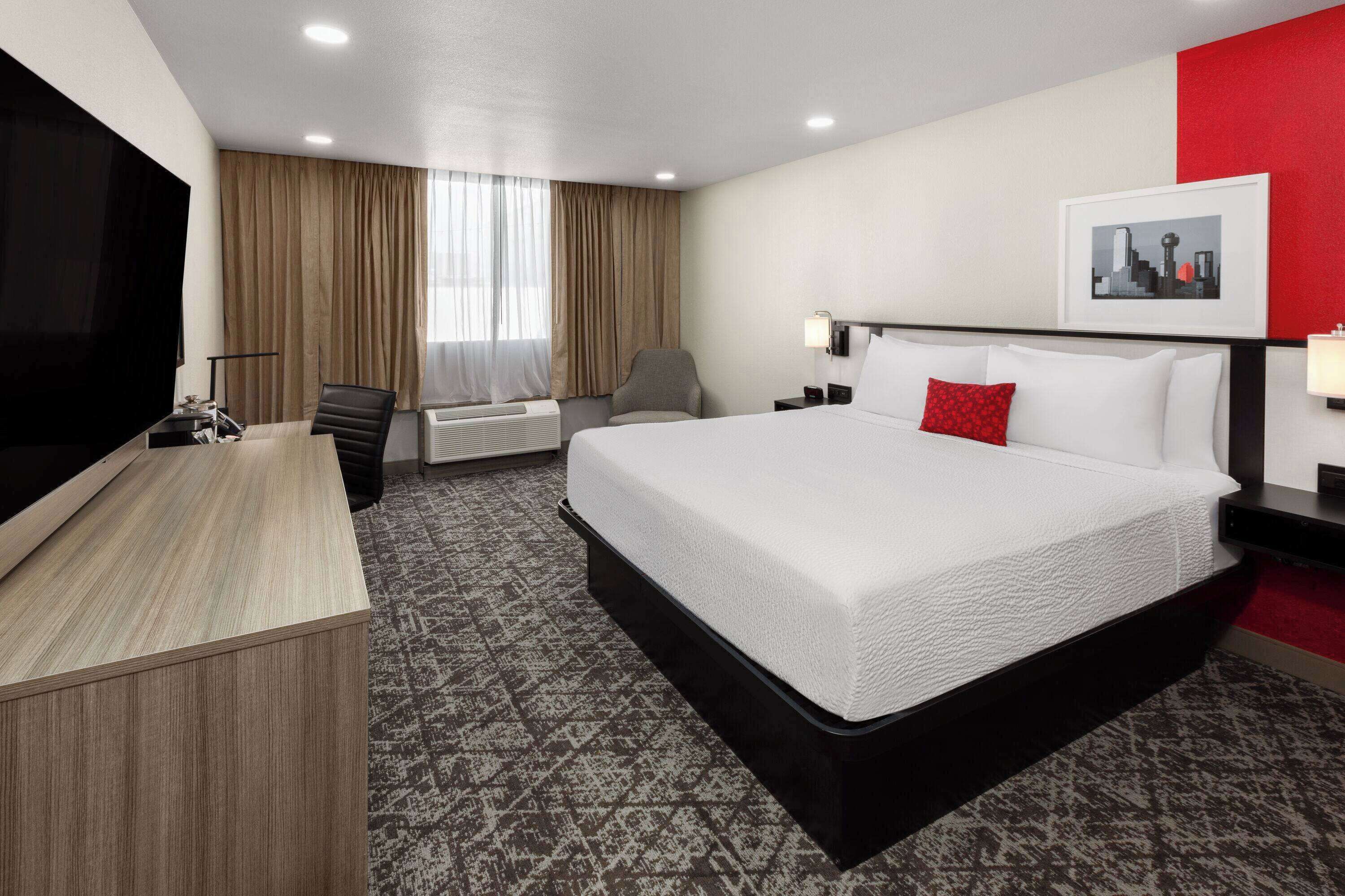 A hotel room with a large bed, a desk, a chair, a TV, and a cityscape artwork on the wall.
