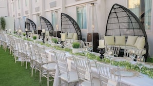 A long dining table with clear chairs, set with white tablecloths and glassware, flanked by black cushioned seating and greenery.