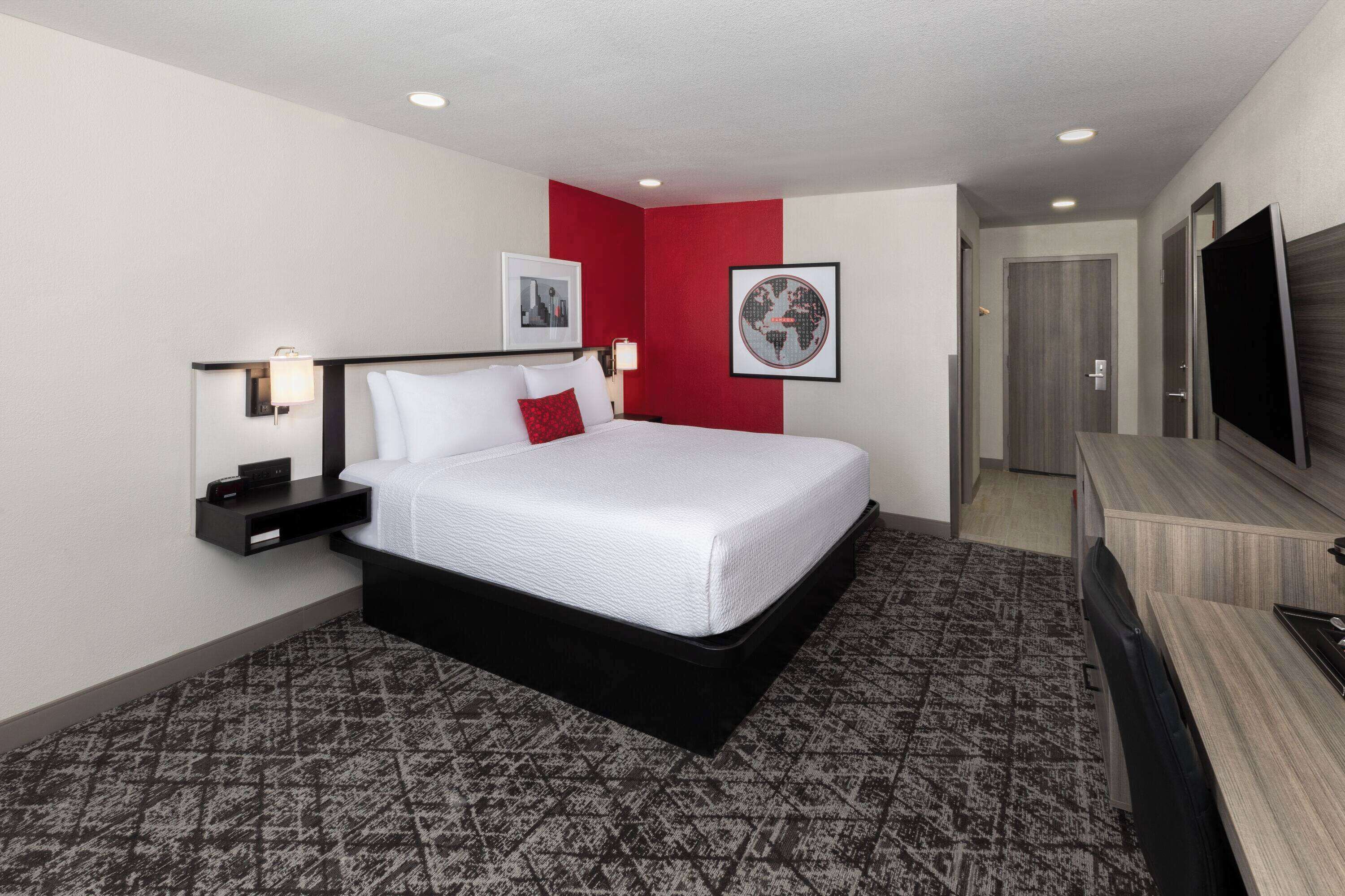 A hotel room with a large bed, a desk, a TV, and a red accent wall.