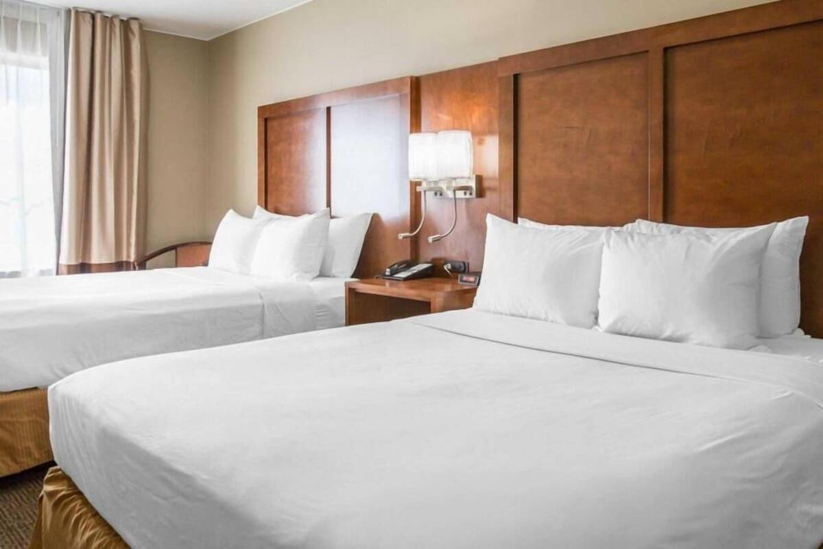 A hotel room with two beds, a wooden headboard, a bedside table with a lamp, and a window with curtains.