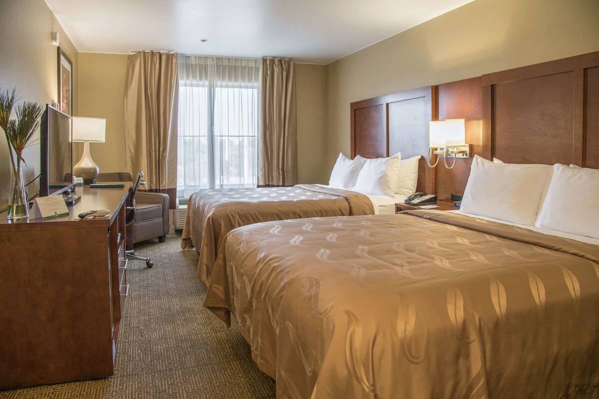 A hotel room with two beds, a desk, a chair, a lamp, and a window with curtains.