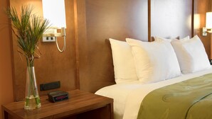 A hotel room with a wooden nightstand, a vase with green plants, a digital clock, and a bed with white bedding and a green throw blanket. - Quality Inn Merced Gateway to Yosemite (Merced)