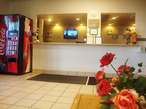 A reception area with a Coca-Cola vending machine, a television screen, and a wooden table with a vase of roses. - Topeka Plaza Inn (Topeka)