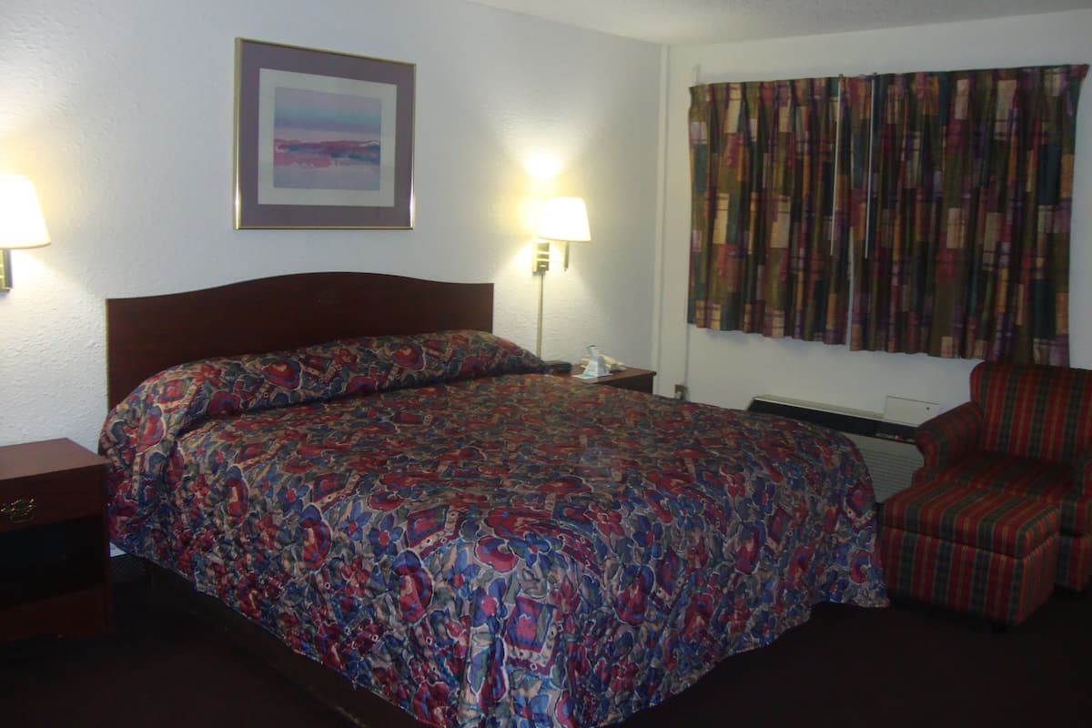 A hotel room with a large bed, two bedside lamps, a framed picture on the wall, a window with curtains, and a chair.