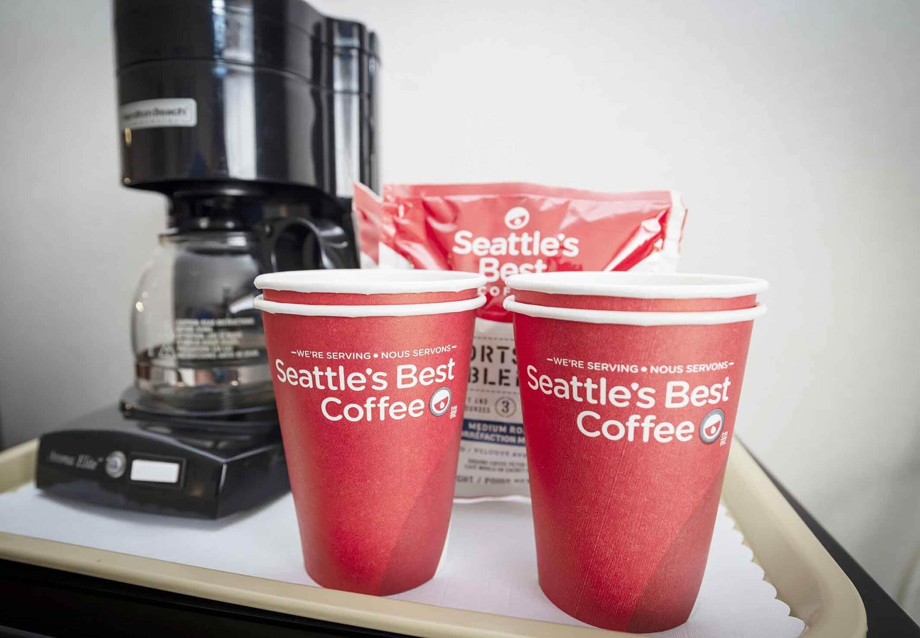 Two red cups labeled "Seattle's Best Coffee" on a tray with a coffee maker and a bag of coffee in the background.