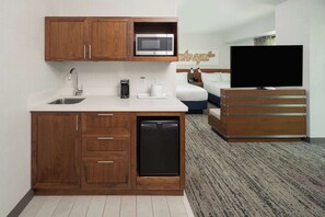 Habitación de hotel compacta con kitchenette, que incluye microondas, cafetera y lavavajillas. Hay un televisor y una cama con cabecero.
