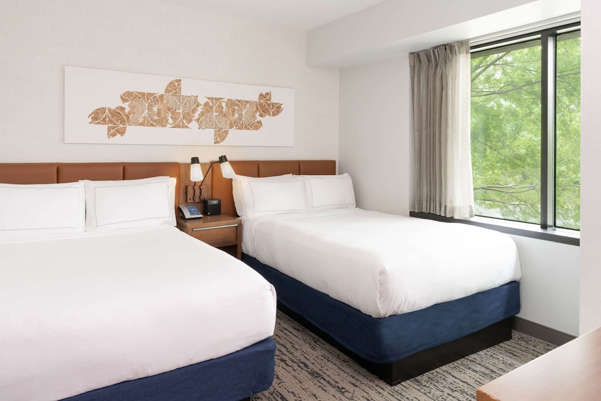 A hotel room with two beds, a nightstand with a lamp, a window with curtains, and a wall-mounted artwork.