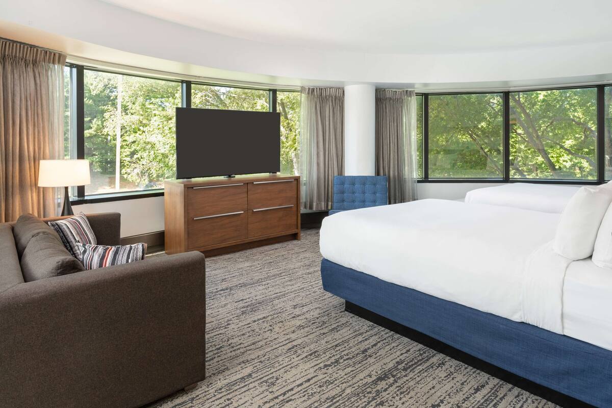 A hotel room with a large bed, a sofa, a TV on a wooden cabinet, and large windows with curtains.