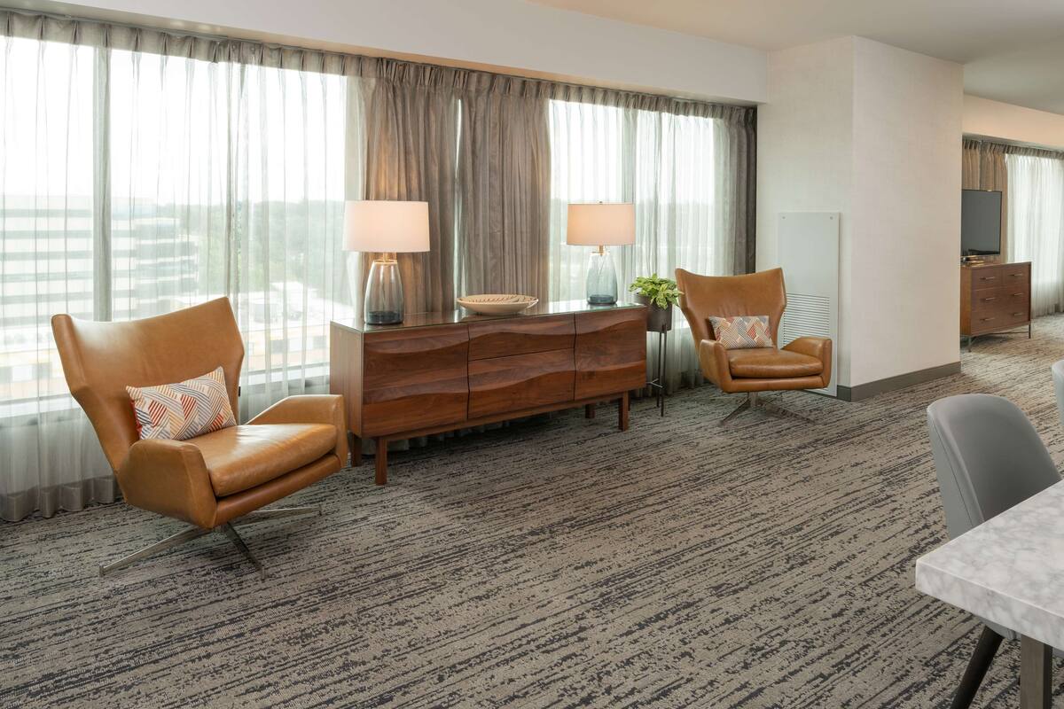 A hotel room with a large window, a wooden dresser, two leather armchairs, a dining table, and a TV on a stand.