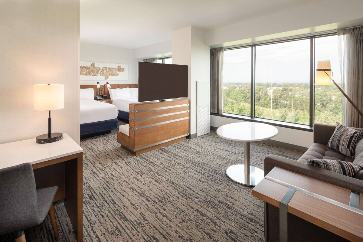 A hotel room with a bed, a TV, a desk, a sofa, and a large window offering a city view.