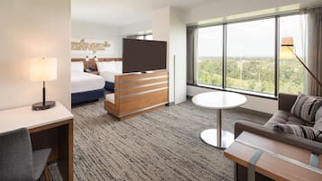 A hotel room with a bed, a TV, a desk, a sofa, and a large window offering a city view.