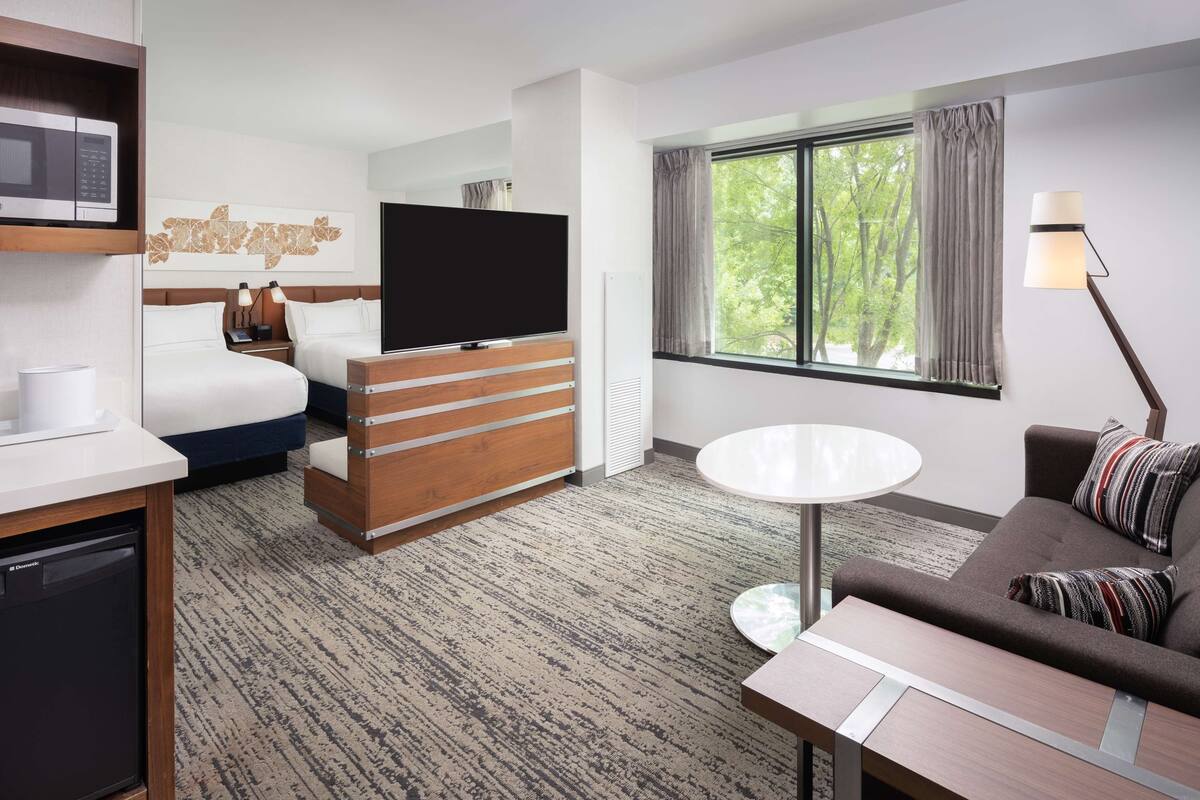 A hotel room with a bed, a TV, a desk, a dining table, and a window with a view of trees.