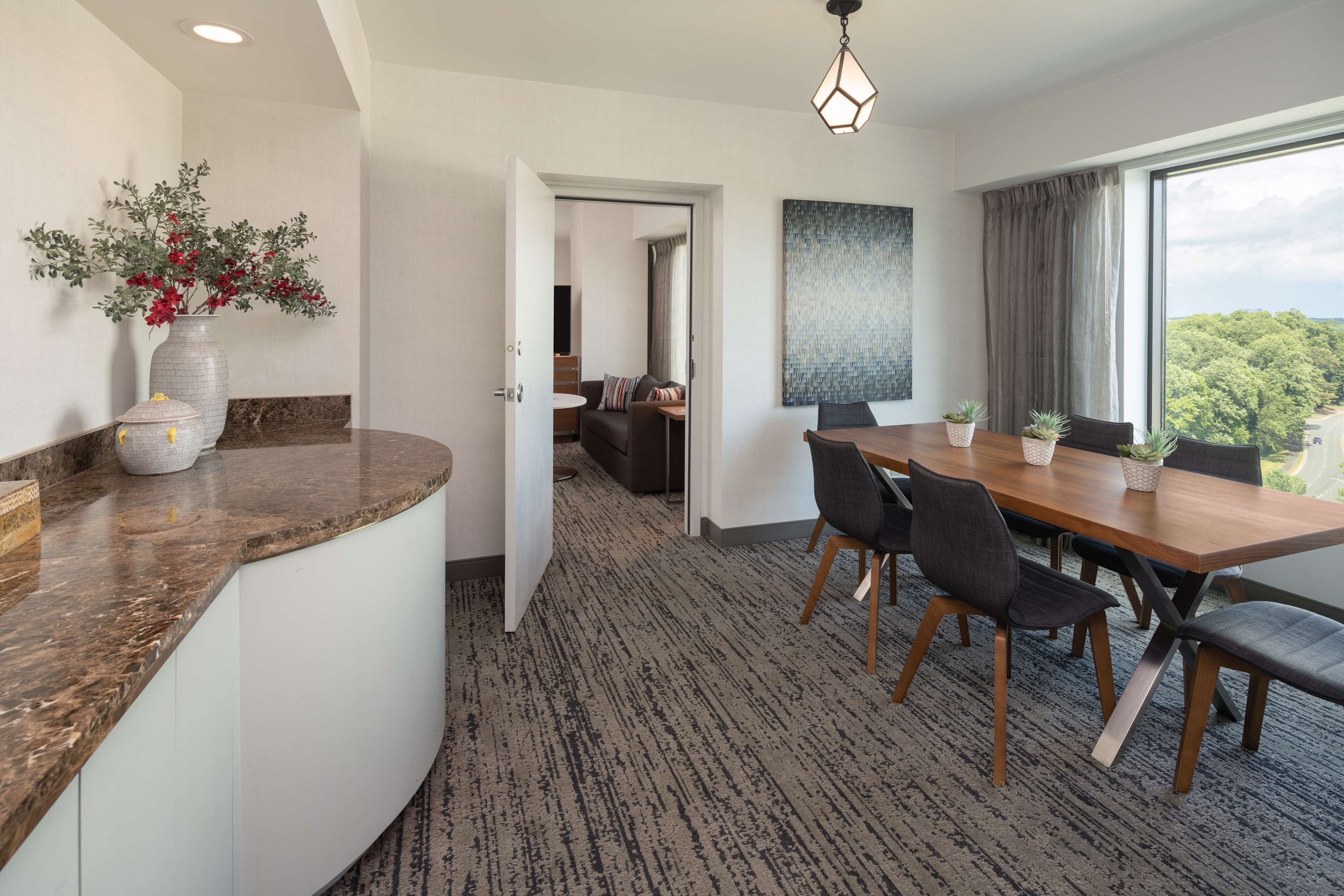 Una habitación de hotel moderna con una encimera de mármol curvada, un área de comedor con una mesa y sillas de madera, un ventanal con vistas a árboles y un pasillo que conduce a otra habitación.