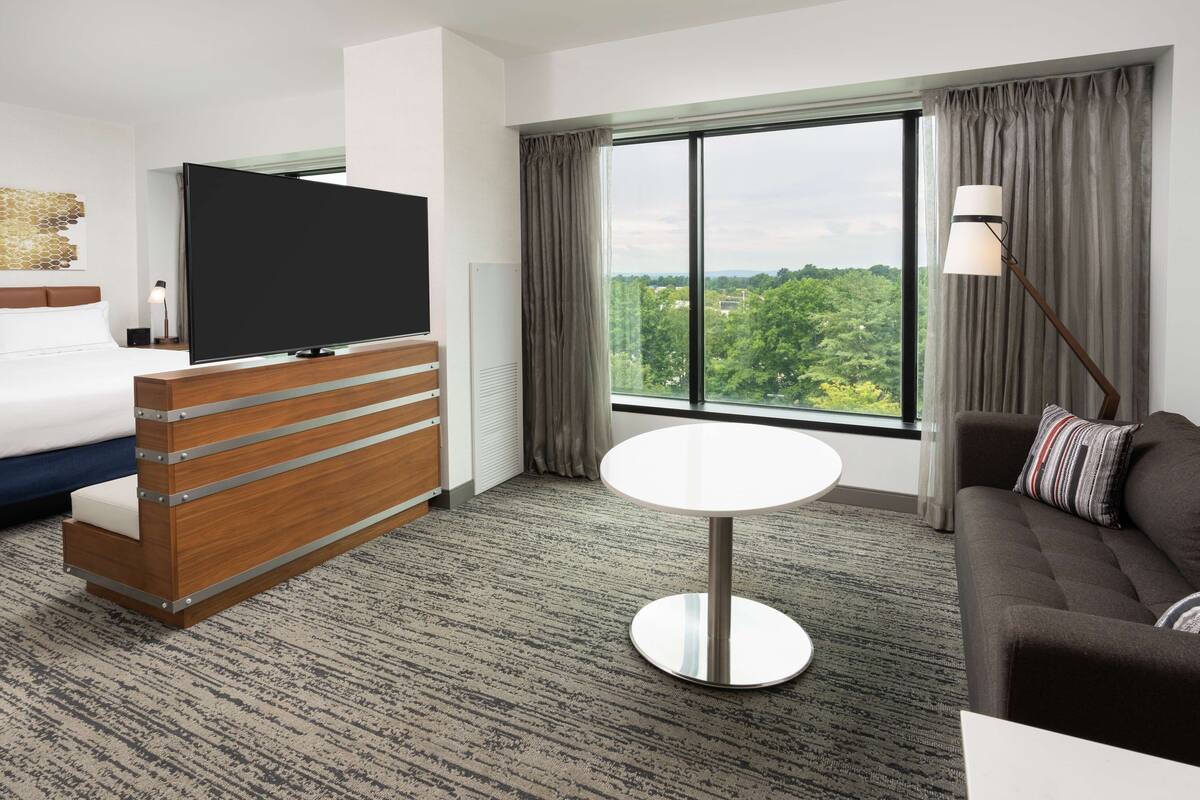 A hotel room with a bed, a TV, a desk, a sofa, and a large window with a view.
