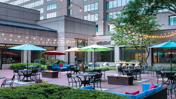 An outdoor patio area with multiple tables and chairs, surrounded by greenery and umbrellas. - Hilton Fairfax (Fairfax)