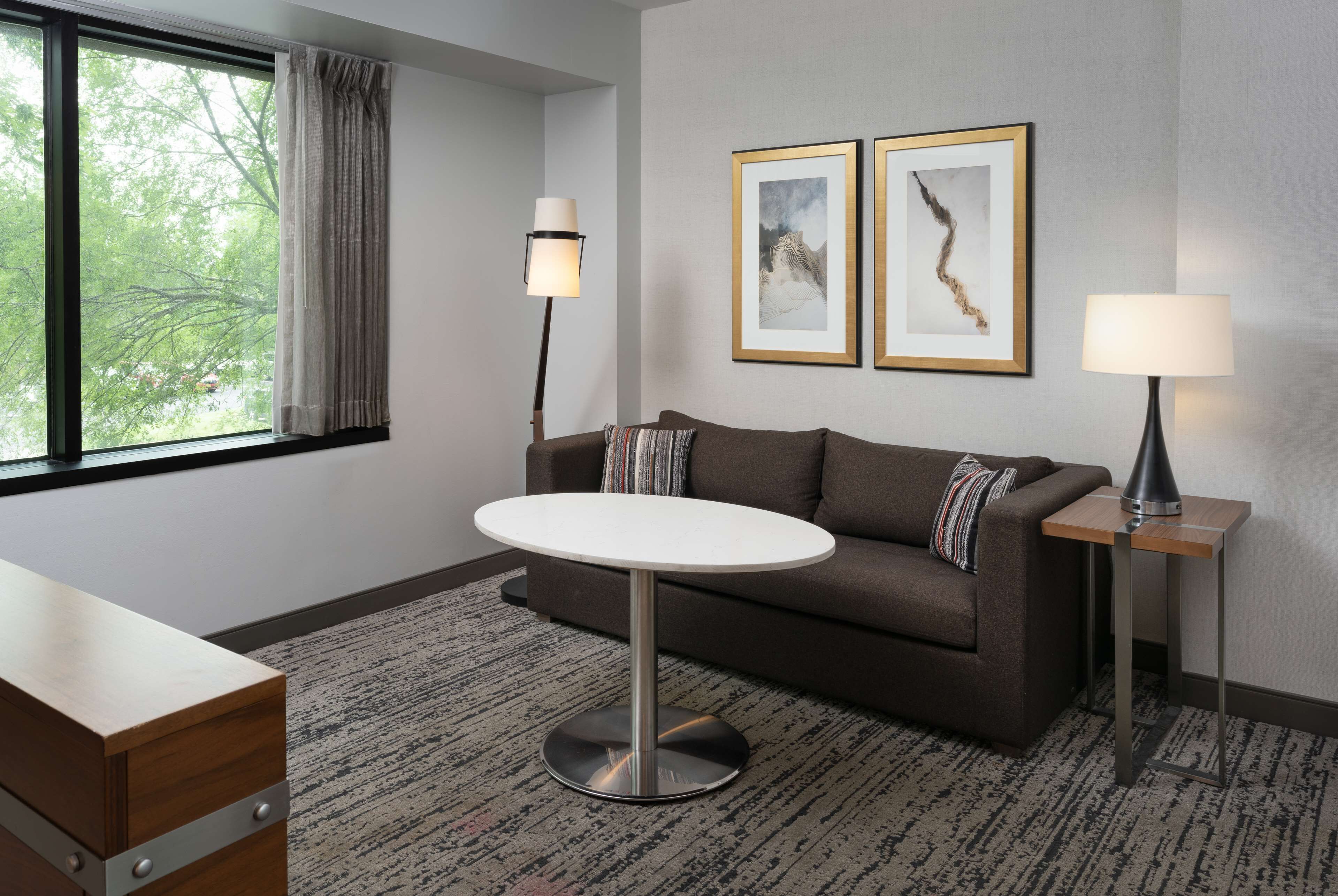 A modern hotel room with a round table, a sofa, two framed artworks, and a view of trees through a window.
