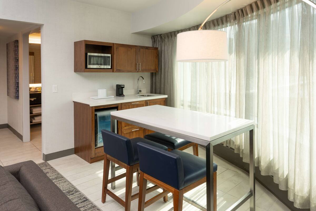 A modern kitchen with wooden cabinets, a white countertop, and a built-in microwave. There is a dining area with a white table and two blue chairs.