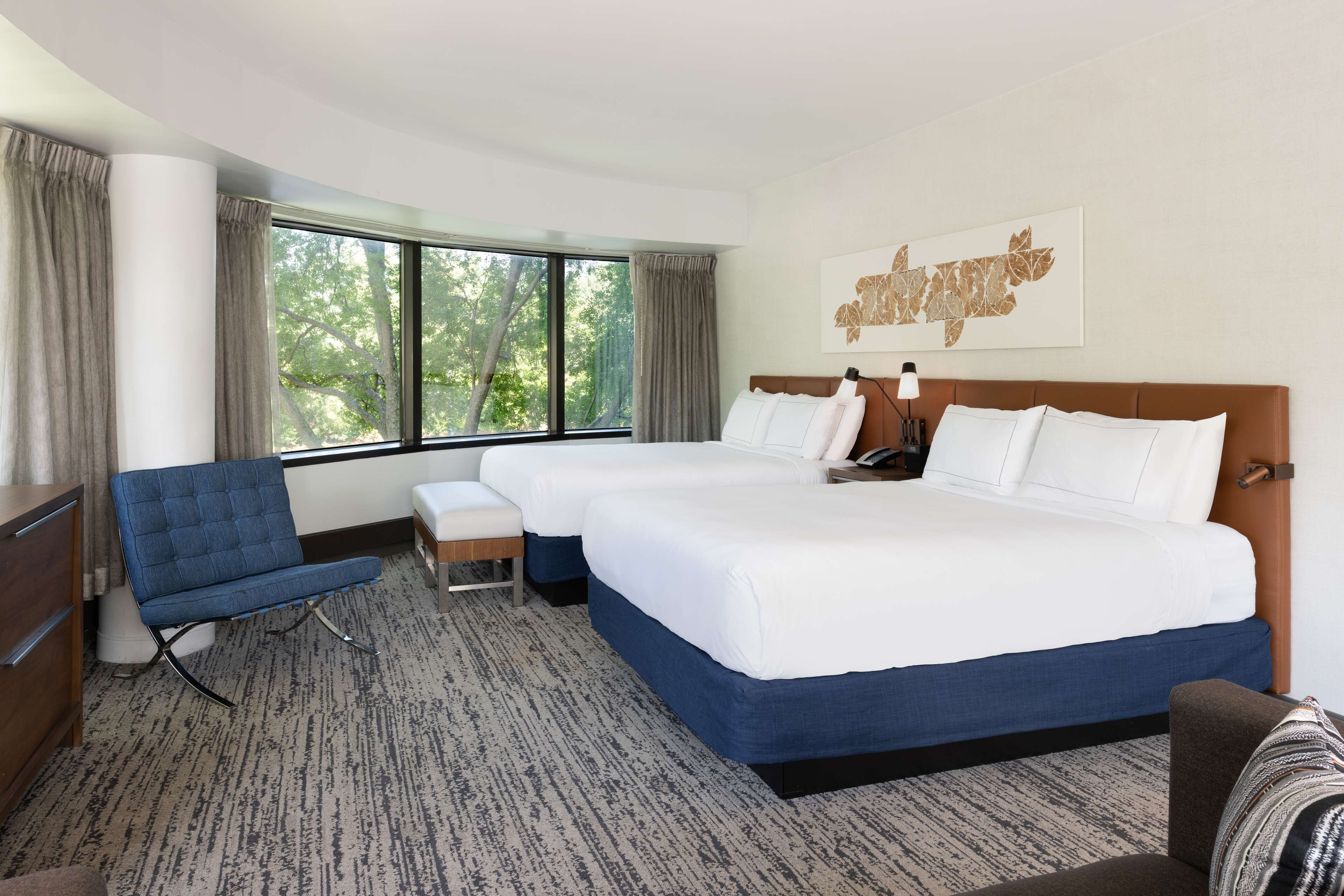 Une chambre d’hôtel avec deux lits, un fauteuil bleu, une petite table et une grande fenêtre donnant sur un paysage arboré.