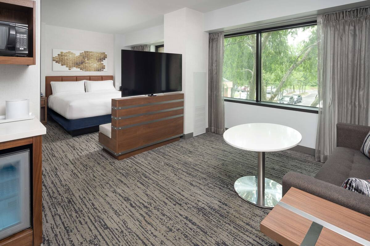 A hotel room with a bed, a TV, a dresser, a round table, and a window with a view of trees.