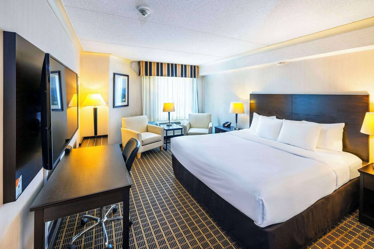 A hotel room with a large bed, a desk with a flat-screen TV, two armchairs, a small table, and two bedside lamps.