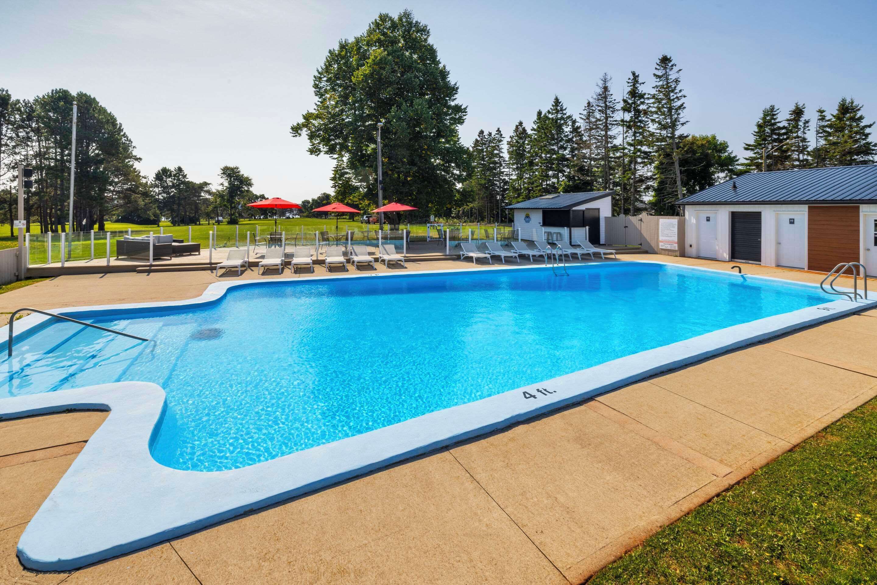 A rectangular swimming pool with a 4 ft. depth marker, surrounded by lounge chairs and umbrellas, with a covered area and a building in the background.