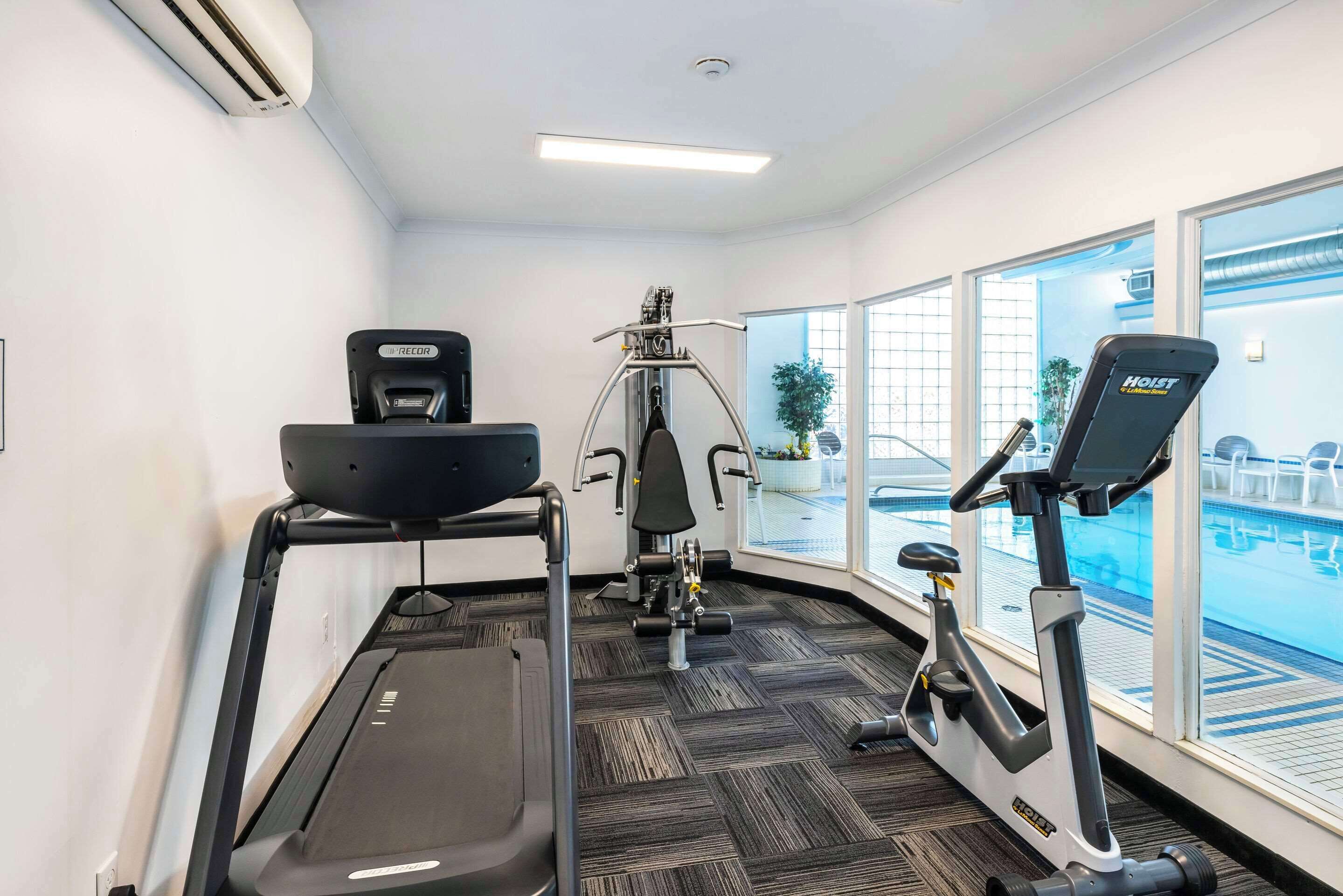A gym with a treadmill, elliptical, and weight machine, large windows, and a view of a pool.