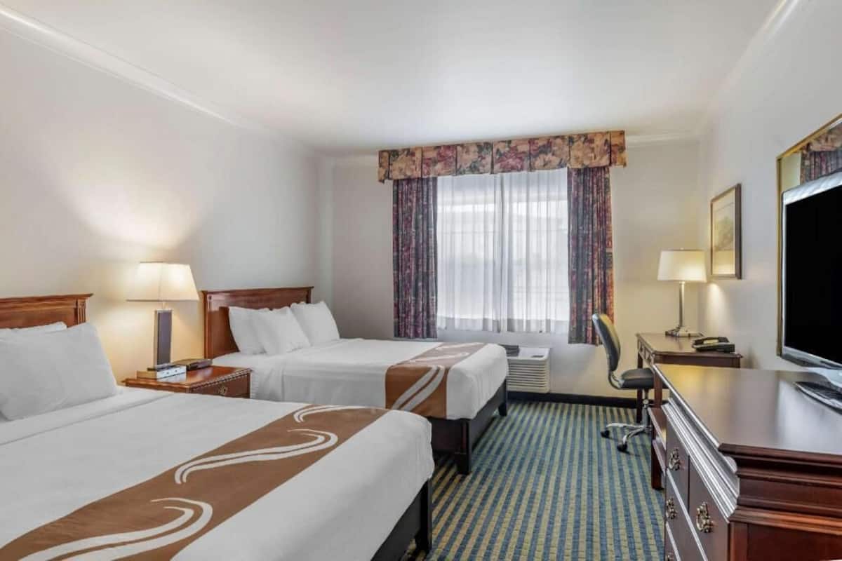 A hotel room with two beds, a desk, a television, and a window with curtains.