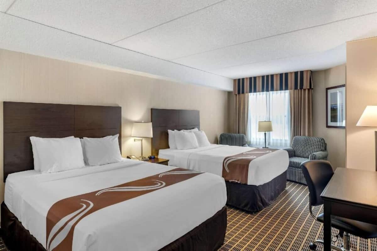 A hotel room with two beds, a desk, a chair, a lamp, and a window with curtains.