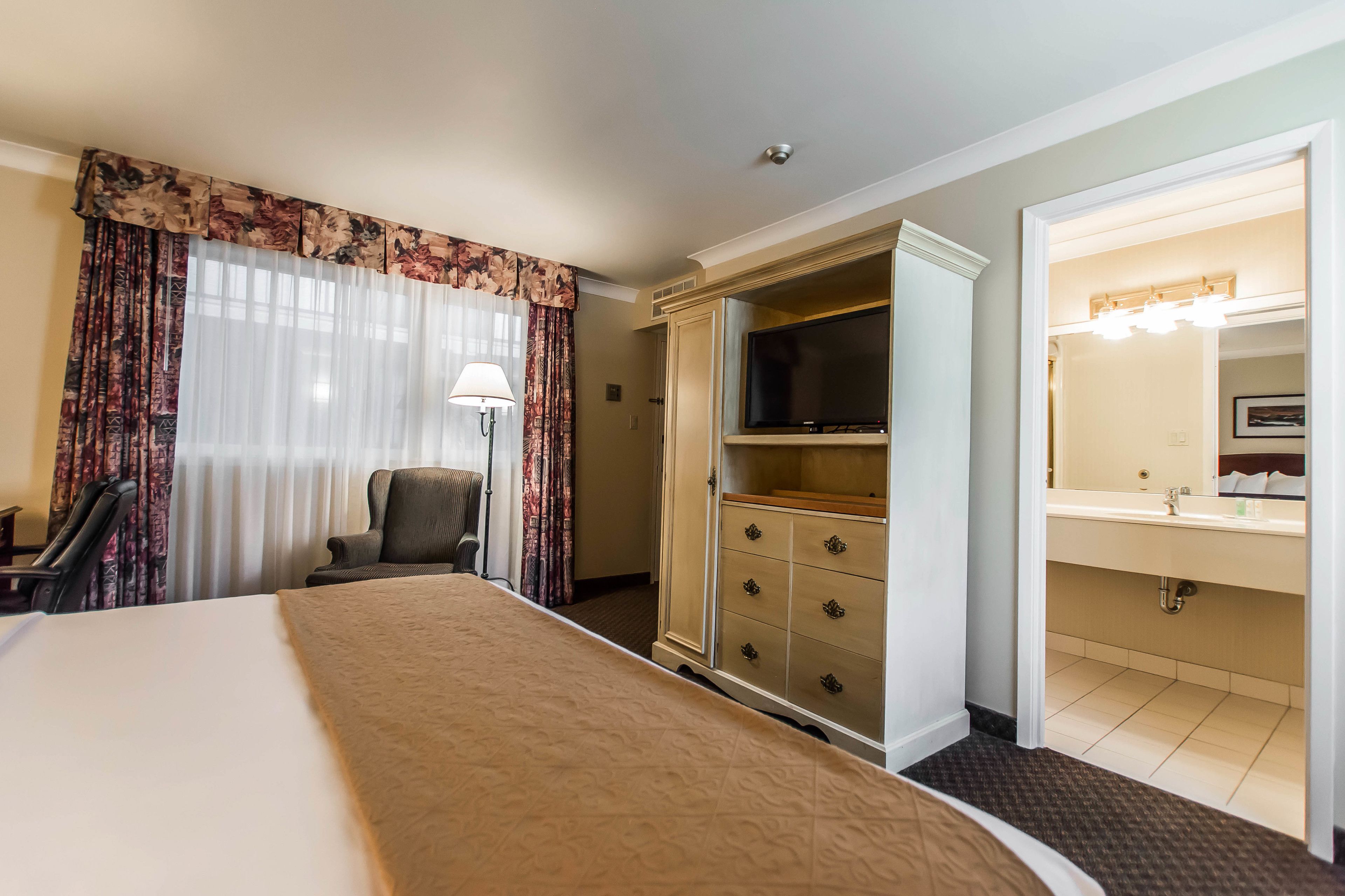 A hotel room with a large bed, a wardrobe, a television, a desk, and a bathroom with a sink and mirror.