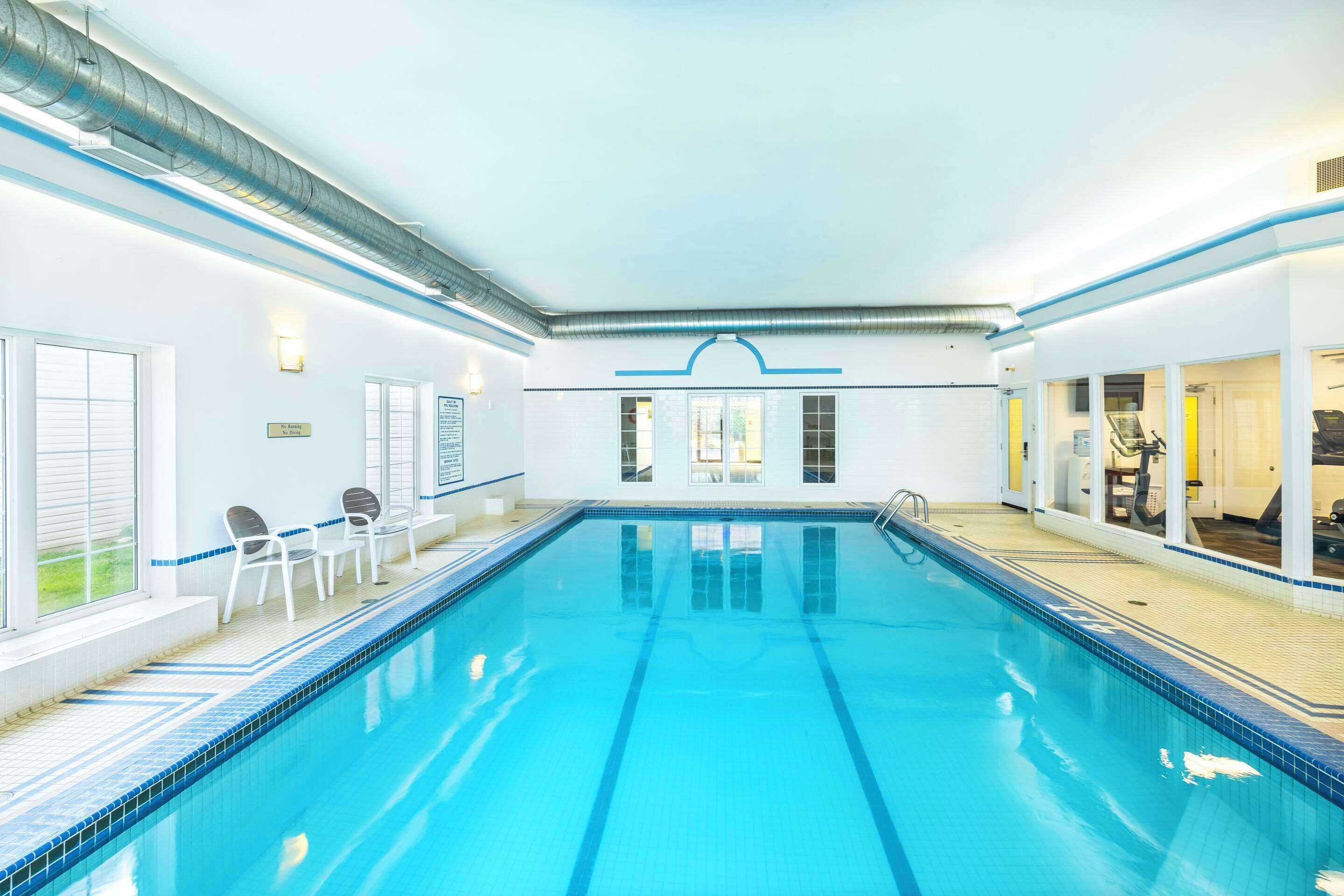 Una piscina interior con agua azul cristalina, rodeada de paredes blancas y un suelo de baldosas. Se observan ventanas en un lateral y una puerta que da a otra habitación.
