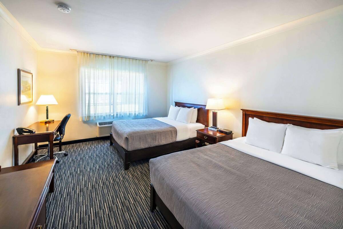 A hotel room with two beds, a desk, a chair, a lamp, and a window with curtains.