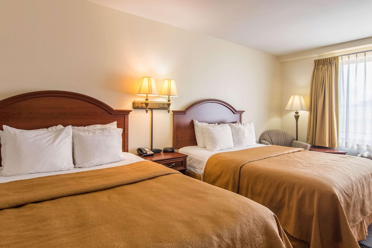 A hotel room with two beds, each with a wooden headboard, white bedding, and a brown blanket. There are bedside lamps, a nightstand with a phone, and a window with curtains.