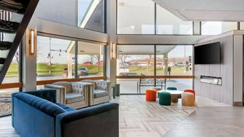 A spacious lobby with a modern design, featuring a large seating area with sofas and ottomans, a flat-screen TV mounted on the wall, and large windows offering a view of the parking area.