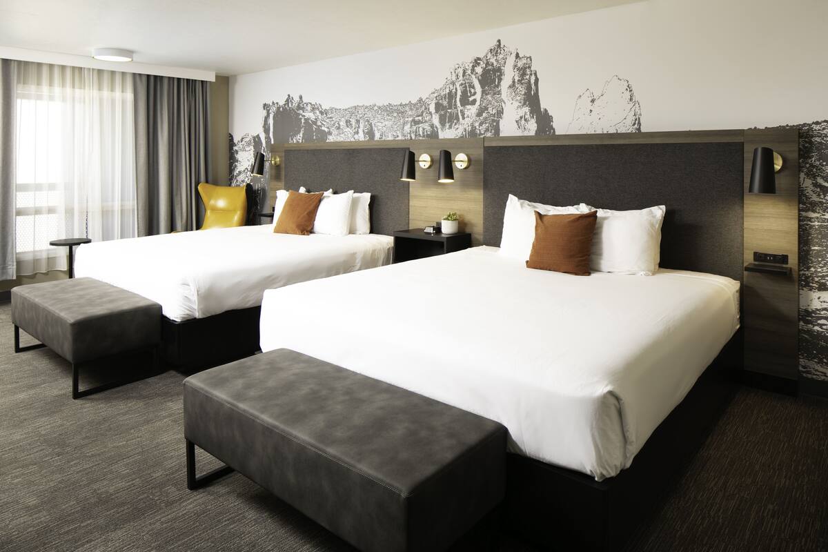 Quarto de hotel com duas camas, bancos estofados em cinza, uma mesinha e uma televisão fixada na parede.
