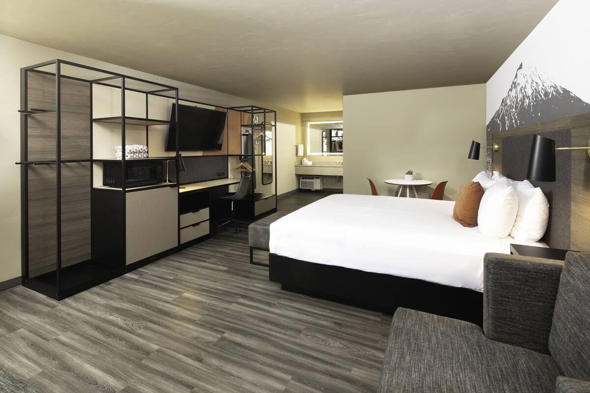 Quarto de hotel moderno com uma cama grande, uma escrivaninha com cadeira, uma TV e uma mesinha com outra cadeira.