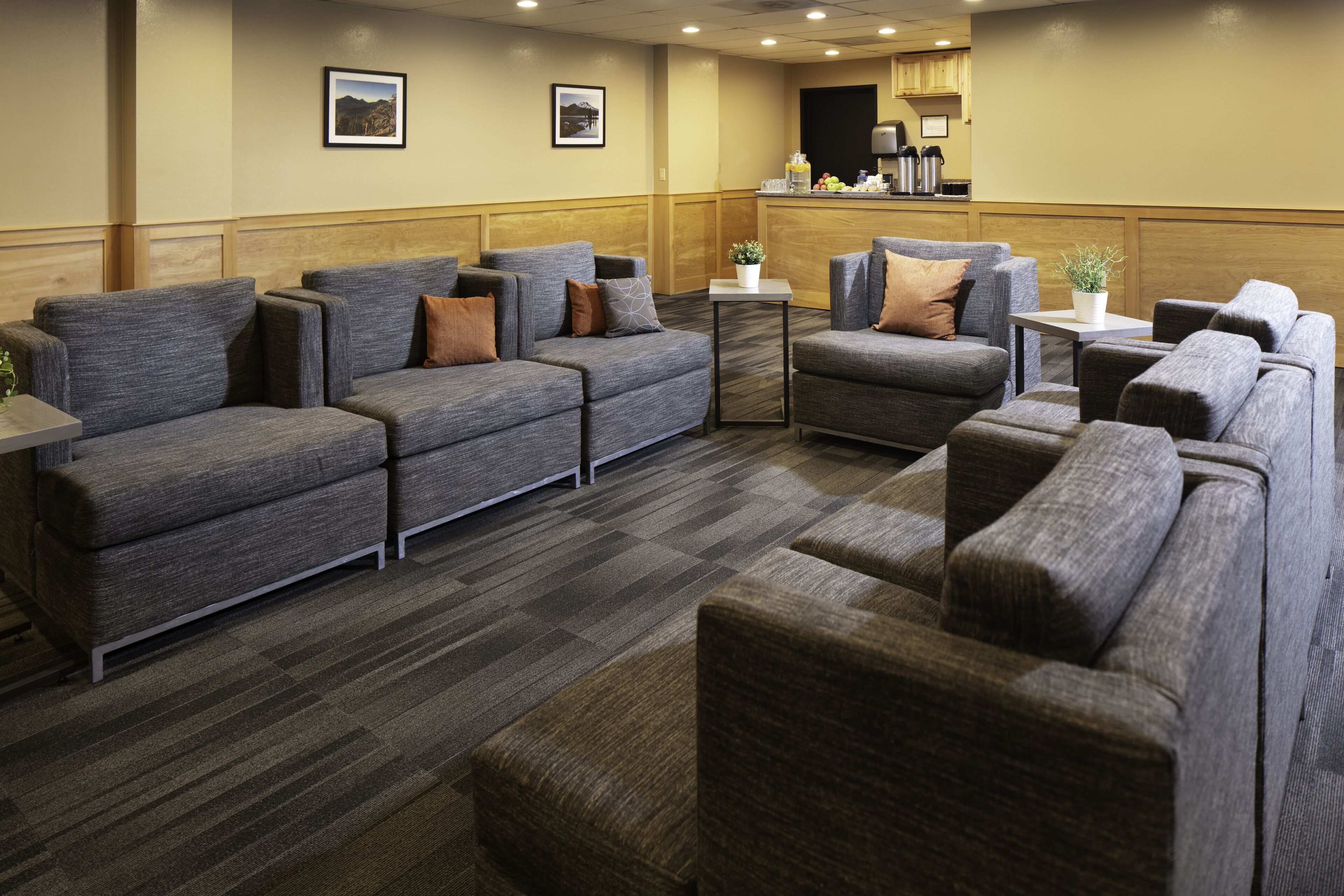 A hotel lobby with grey sofas and armchairs, a small table with a plant, and a kitchen area in the background.