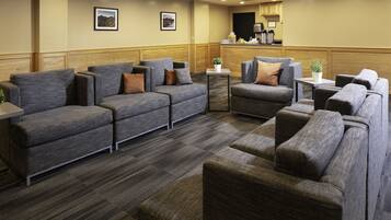 A hotel lobby with grey sofas and armchairs, a small table with a plant, and a kitchen area in the background.