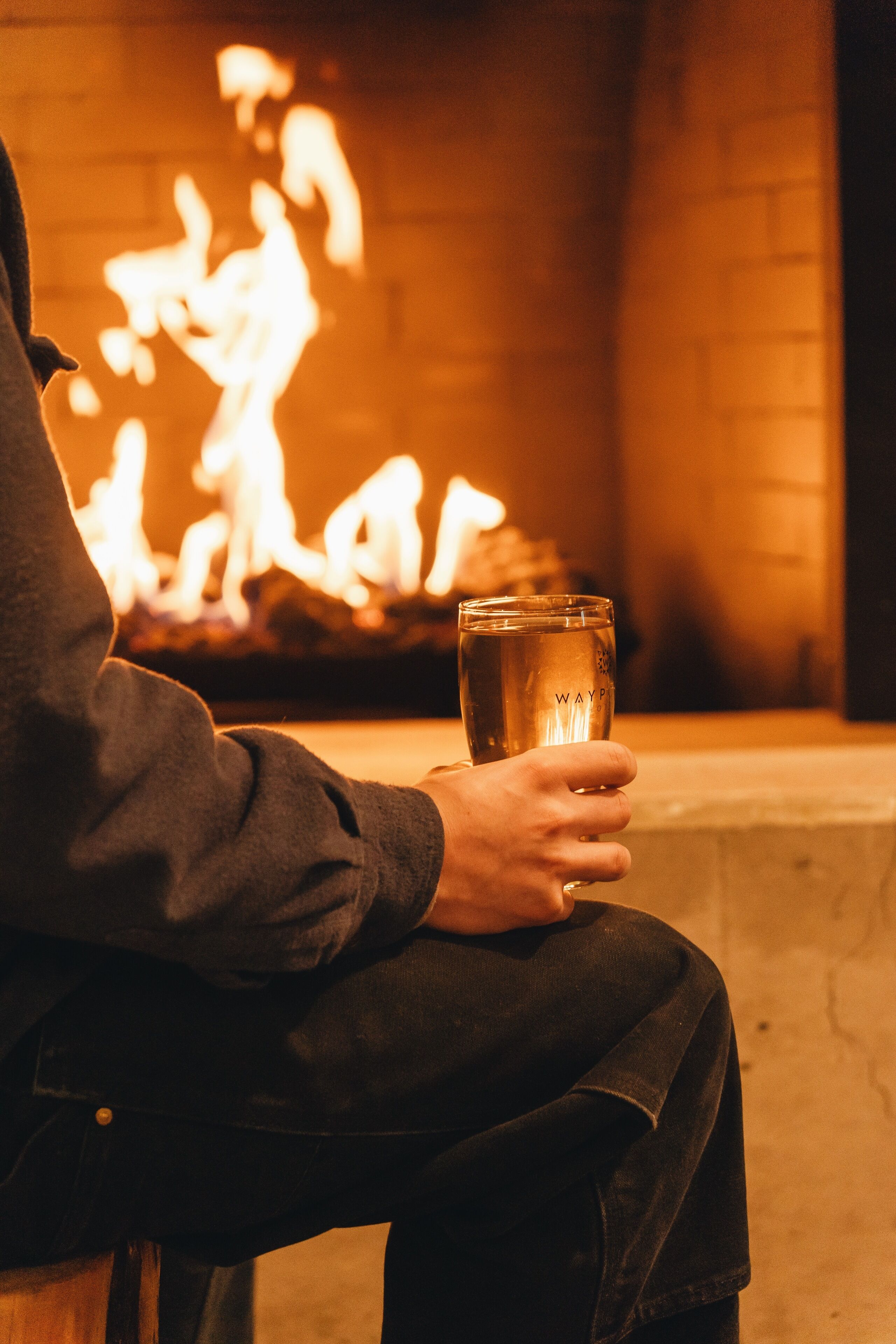 Une personne tenant un verre sur lequel est inscrit le mot « WAYPA », assise devant une cheminée.