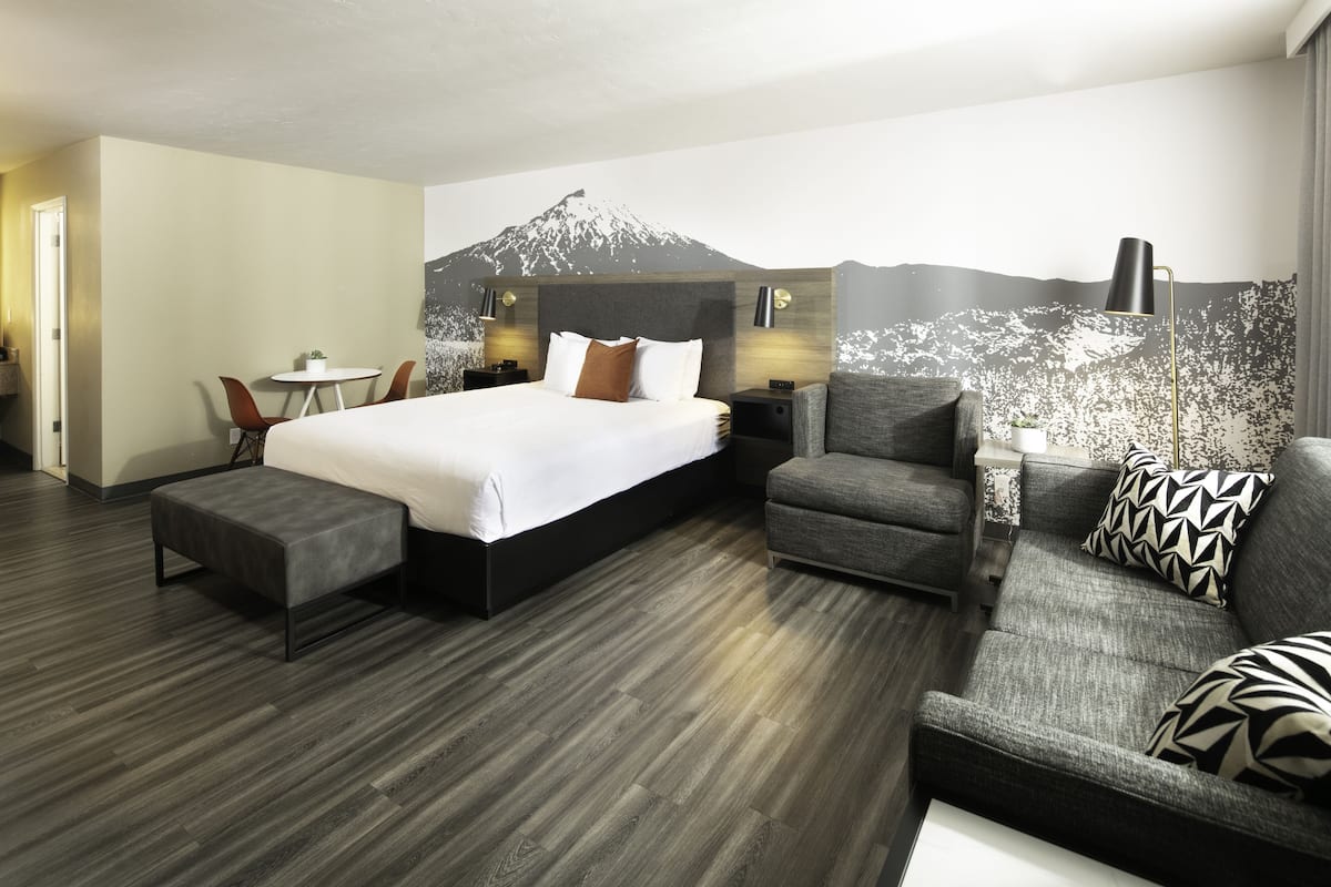 Quarto de hotel moderno com uma cama grande, um sofá cinzento, uma mesinha e uma cadeira.