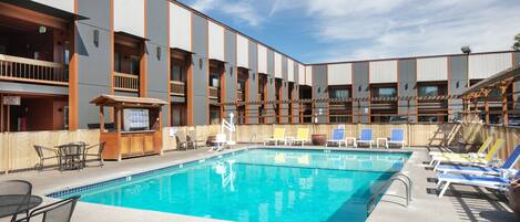 A swimming pool with lounge chairs and a wooden deck area, surrounded by a multi-story building with balconies.