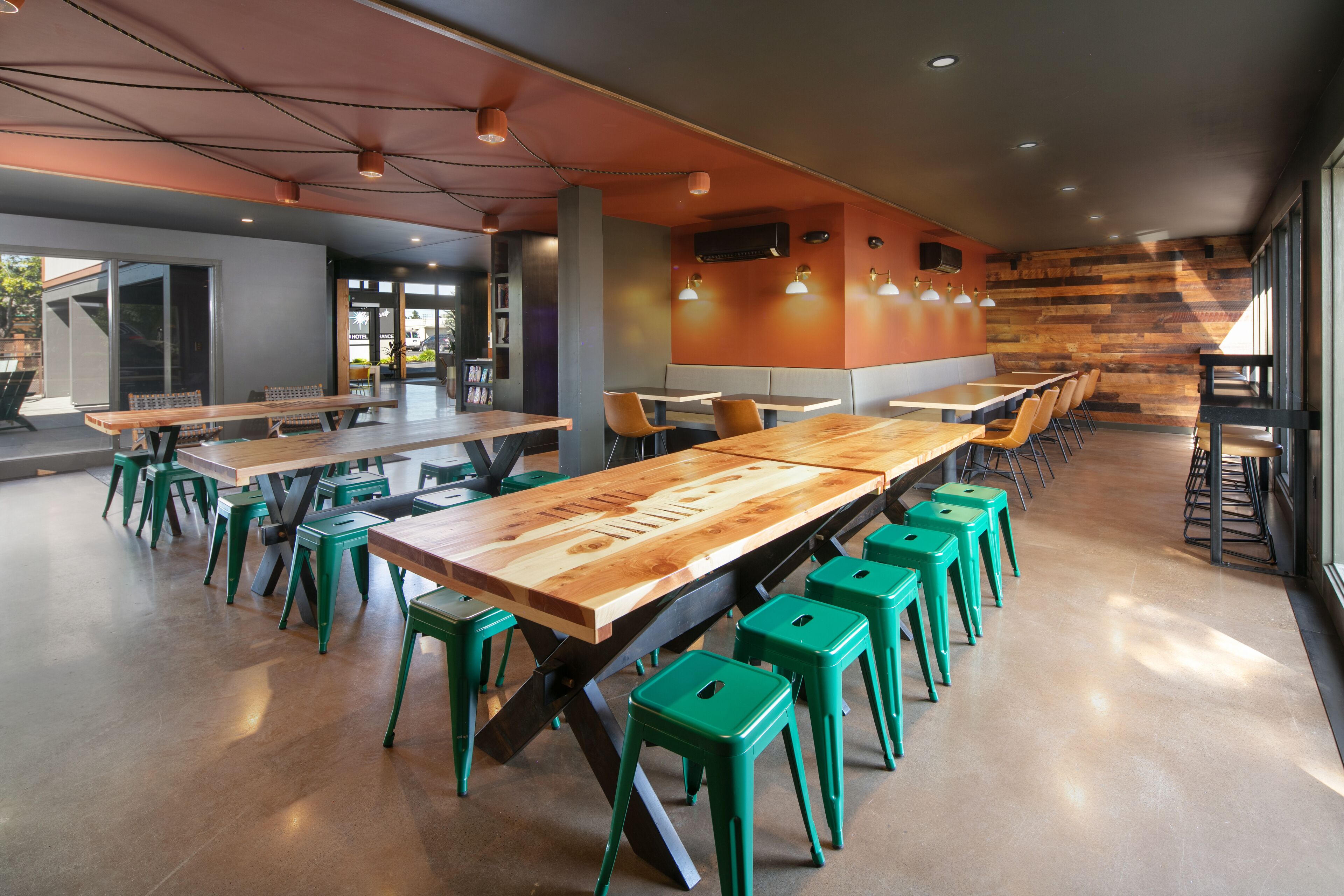 A modern dining area with long wooden tables and green stools, featuring a mix of natural and artificial lighting.