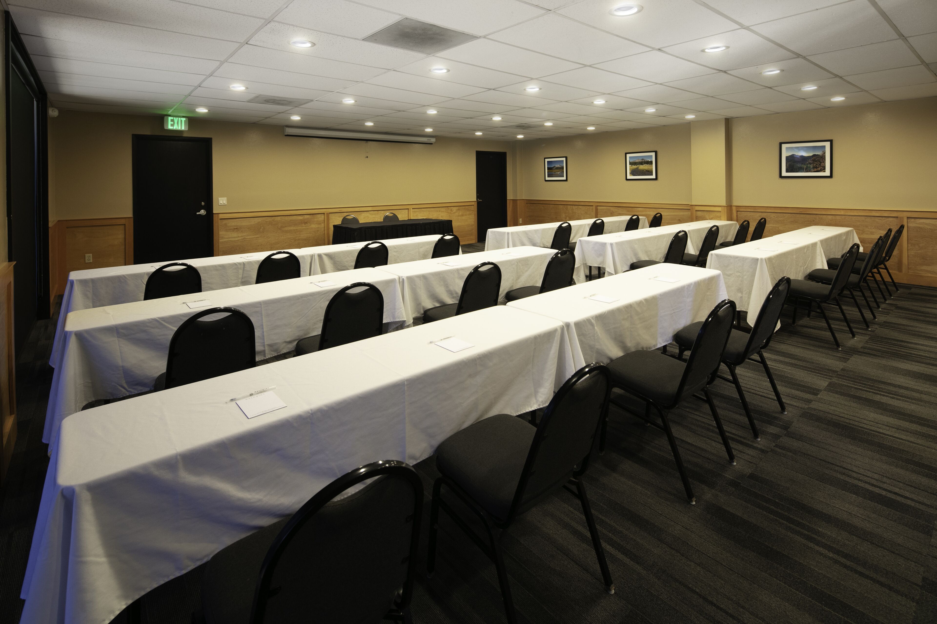 A conference room with multiple tables and chairs, an exit door, and framed pictures on the walls.